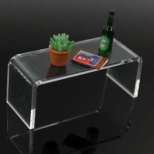 Può includere: Un tavolino in acrilico trasparente con angoli arrotondati. Sul tavolo ci sono una piccola pianta grassa in vaso, un libro con copertina rossa e blu e una bottiglia di birra Heineken verde. Il tavolo è su uno sfondo nero.