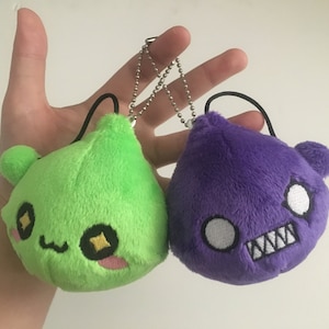 May include: Two plush keychain toys. One is green with a cute face and the other is purple with an angry face.