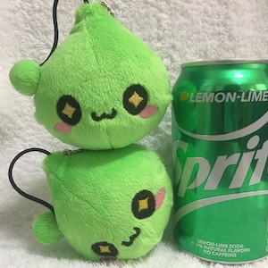 May include: A green plush toy with a smiling face and large eyes. The toy is shaped like a teardrop and has a small black string attached to the top. The toy is sitting next to a can of Sprite Lemon-Lime soda.