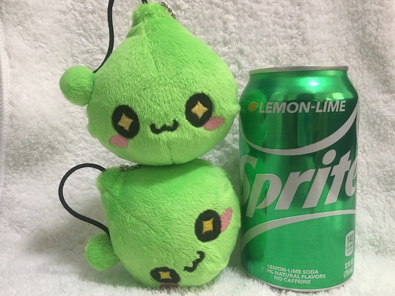 May include: A green plush toy with a smiling face and large eyes. The toy is shaped like a teardrop and has a small black string attached to the top. The toy is sitting next to a can of Sprite Lemon-Lime soda.