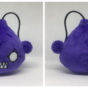 May include: Purple plush toy shaped like a teardrop with a black string loop on top. The toy has a white face with two large eyes and a toothy grin.