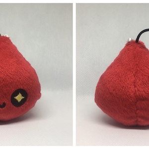 May include: Red plush toy shaped like a teardrop with a black string loop. The toy has a cute face with black eyes and a small mouth. The eyes have gold glitter.