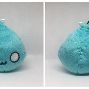 May include: A teal blue plush toy shaped like a teardrop with a black elastic loop on top. The toy has a stitched-on face with white eyes and a small mouth.