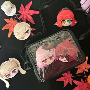 Könnte beinhalten: Eine schwarze, rechteckige Umhängetasche mit einer durchsichtigen Frontpartie, die zwei Anime-Charakterköpfe zeigt. Um die Tasche herum liegen mehrere kleine Anstecknadeln, eine weiße Würfelfigur, eine türkisfarbene Kreatur und rote Ahornblätter.
