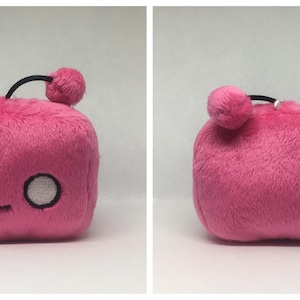 May include: A pink plush toy shaped like a cube with a black antenna and a round pom-pom on top. The toy has a simple embroidered face with two white eyes and a small mouth.