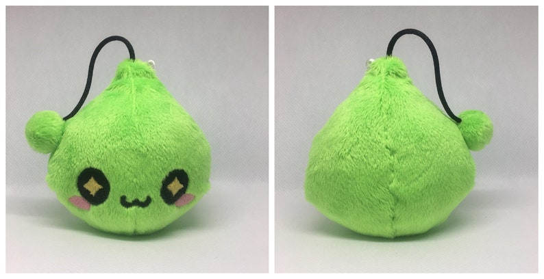 May include: A green plush toy shaped like a teardrop with a cute face and sparkly eyes. The toy has a black string attached to the top and a small green pom-pom on the end of the string.