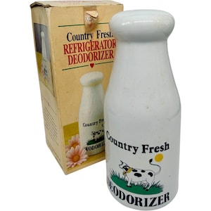 Puede incluir: Un desodorizante para refrigerador de cerámica blanca con forma de botella de leche, con una ilustración de una vaca en blanco y negro, hierba verde y el texto Country Fresh DEODORIZER, junto a su caja de cartón original con gráficos a juego.