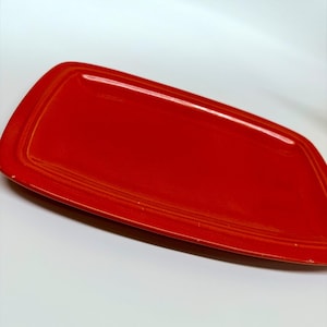 May include: A rectangular ceramic serving tray in a vibrant, glossy red finish. The tray features a smooth, flat central surface surrounded by a slightly raised, rounded border that frames the entire perimeter, creating a clean, simple design.