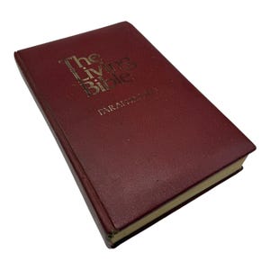 Peut inclure: Un livre de couleur marron intitulé "The Living Bible" avec des lettres dorées. Le livre est une édition à couverture rigide avec un aspect légèrement usé. Le texte "PARAPHRASED" est imprimé en dessous du titre.