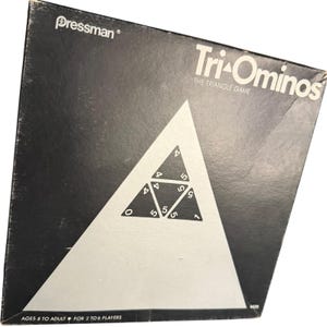 May include: A black square board game box for Tri-Ominos The Triangle Game by Pressman. The cover features a large white triangle containing smaller black triangular tiles with white numbers 0, 1, 4, and 5. Text includes Ages 8 to Adult.