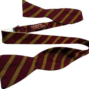 May include: A burgundy bow tie featuring diagonal stripes in yellow and green. The fabric has a ribbed texture. A label on the inner strap reads CHAPS by RALPH LAUREN ALL SILK. The bow tie includes an adjustable metal hook and eye closure.