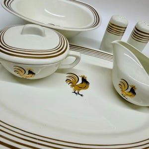 May include: A cream-coloured ceramic dinnerware set featuring a brown rooster graphic and brown concentric line accents. The collection includes a large oval platter, a lidded bowl with a handle, a gravy boat, and two cylindrical salt and pepper shakers.