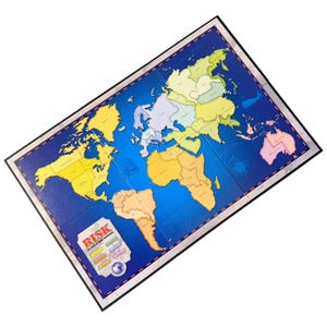 May include: A board game map of the world, featuring continents in various colors against a blue ocean background. The game's title, "RISK", is displayed on a blue emblem in the lower left corner. The board is rectangular with a black border.