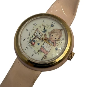 May include: Vintage Picco watch with a light pink band and a gold-tone bezel. The watch face features a cartoon illustration of a child and the text "Put on a Happy Face!". The watch is a nostalgic item.