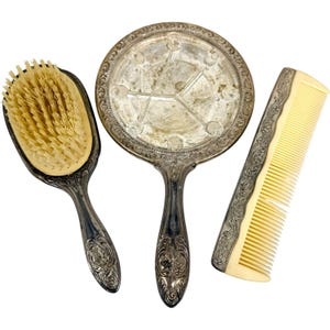 Pode incluir: Um conjunto de toucador composto por uma escova de cabelo com cerdas claras, um espelho de mão redondo com moldura prateada manchada e um pente retangular com corpo de plástico creme e borda de cabo de metal prateado ornamentado.