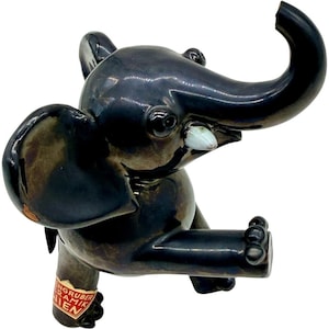 May include: A ceramic elephant figurine in a glossy dark gray finish. The elephant has large ears, a raised trunk, and a light blue tusk. A label with red text is attached to the front leg. The figurine is a decorative item.