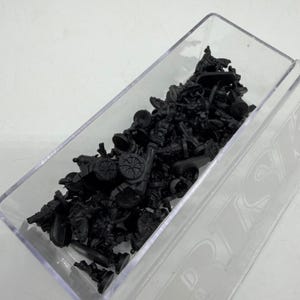 Puede incluir: Un recipiente de plástico transparente lleno de numerosas figuras en miniatura negras. Las figuras parecen ser de varias formas y tamaños, posiblemente representando soldados, cañones y otros elementos relacionados con la guerra. El recipiente está inclinado y el contenido está densamente empaquetado.