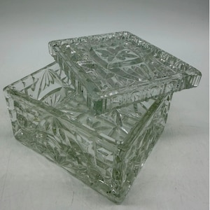 May include: A clear, square glass box with a removable lid. The exterior surfaces of both the box and the lid feature a pressed, faceted geometric pattern with starburst motifs. The glass is transparent and reflects light across its textured edges.