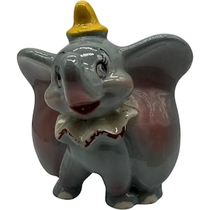 May include: A ceramic figurine of Dumbo the elephant, featuring a gray body, large ears, and a yellow hat. The figurine has a white ruffled collar with red accents and a pink trunk. The figure is standing and smiling.