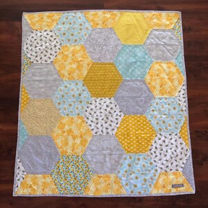May include: A handmade quilt with a honeycomb design. The quilt uses yellow, grey, and light blue fabrics. The hexagonal shapes are arranged in a repeating pattern. The quilt has a light grey border. The quilt's dimensions are approximately 100cm x 100cm.