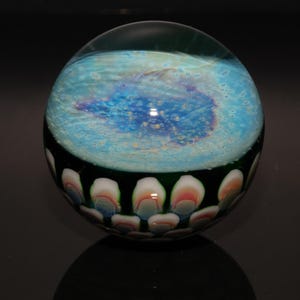 May include: A glass paperweight with a blue and white swirling interior, resembling a galaxy. The bottom edge features a pattern of white, pink, and blue petal-like shapes. The paperweight is spherical and sits on a reflective black surface.