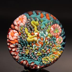 May include: A colorful, round glass art piece with a detailed, abstract design. The artwork features a mix of vibrant colors, including teal, orange, yellow, and red, arranged in a swirling, organic pattern. The glass appears to have a glossy finish.