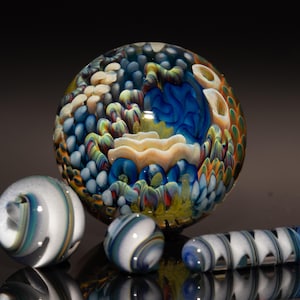 May include: A selection of decorative glass marbles and a glass rod. The large marble has a detailed, swirling design with blue, yellow, and white colours. Smaller marbles and a rod with a spiral pattern are also present.