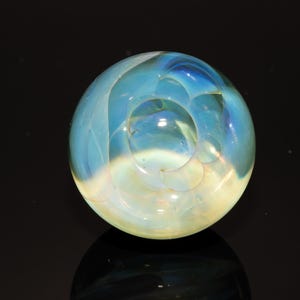 May include: A clear glass sphere with swirling blue and yellow hues. The orb appears to have an internal design, with a central hollow area. The glass is reflective, and the sphere sits on a dark surface.