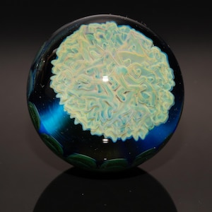 May include: A clear glass marble with a swirling, abstract design. The center features a light yellow and white pattern, surrounded by a deep blue and black background. Green leaf-like shapes are visible around the edge. The marble is round and reflects light.