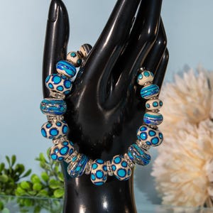 May include: A beaded bracelet featuring a variety of blue and cream-colored beads. The bracelet has a mix of round and cylindrical beads with a silver-colored spacer. The beads have a spotted and striped design.