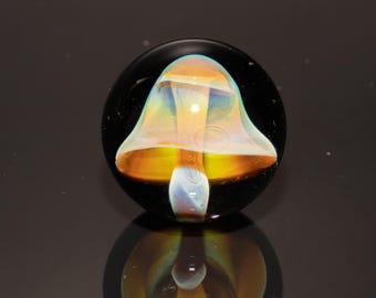 May include: A glass mushroom paperweight with a black base. The mushroom cap is a translucent orange and yellow, with a white stem. The paperweight is round and reflects the mushroom's image below.