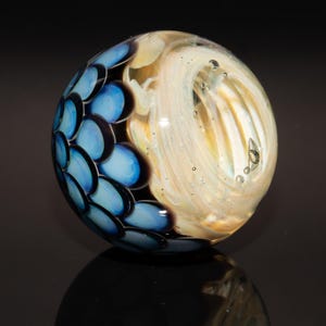 May include: A handcrafted glass marble with a unique design. The marble features a pattern of light blue and dark blue scales on one side, transitioning to a swirling, cream-colored pattern on the other. Small air bubbles are visible within the glass.