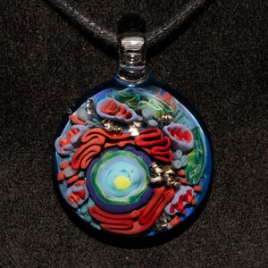 Op de afbeelding: Een ronde glazen hanger met een gedetailleerd, kleurrijk ontwerp. De hanger heeft een blauwe achtergrond met rode, blauwe, groene en gele gebeeldhouwde elementen. Hij hangt aan een zwart koord.