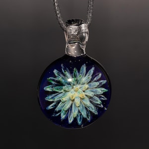May include: A handcrafted glass pendant with a dark blue background and a central floral design in shades of light blue, green, and yellow. The pendant is suspended from a dark gray cord.