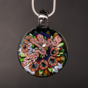 May include: A round, glass pendant with a colorful, abstract design. The pendant features a complex arrangement of orange, blue, pink, and green elements encased in clear glass. It hangs from a silver chain, creating a striking piece of wearable art.