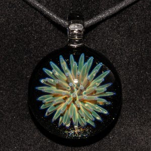 May include: A round, glass pendant with a black background and a central design resembling a stylized flower. The flower has green and blue petals with small, light-colored dots. The pendant hangs from a black cord.