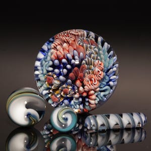 May include: A collection of colorful glass art pieces. A large, round paperweight with red, white, and blue patterns is surrounded by three marbles and a glass rod. The marbles and rod have swirling designs.