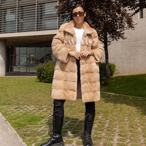 Puede incluir: Un abrigo largo de piel de color marrón claro con cierre de botones. El abrigo tiene un cuello ancho y se lleva sobre una camiseta blanca y pantalones negros. La persona también lleva botas negras con cordones. El abrigo tiene la palabra "SALAMAS".