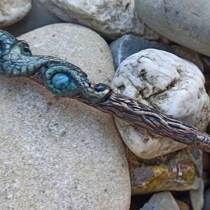May include: A handcrafted pen with a detailed, teal and bronze-colored dragon claw design at the top. The pen's body has a textured, wood-like appearance, and the pen tip is gold-colored.