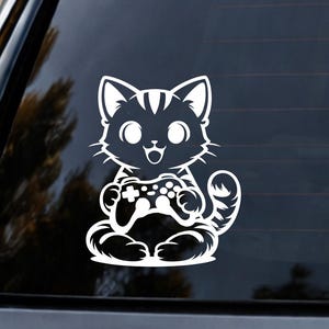 May include: White vinyl decal of a cartoon cat sitting and holding a video game controller. The cat has large eyes and a happy expression. The decal is designed to be applied to a car window or similar surface.