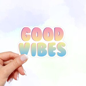 May include: A sticker with the text "GOOD VIBES" in a rainbow gradient font. The sticker has a white outline and is held in a hand.