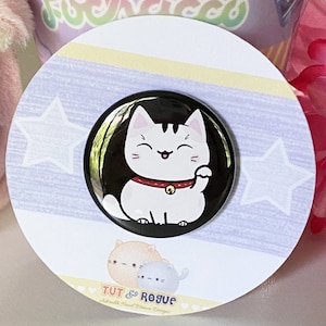 May include: A black and white button featuring a cartoon white cat with black stripes, a red collar with a gold bell, and a raised paw. The cat is smiling and has pink ears. The button is on a white background with a blue and yellow striped border and two white stars.