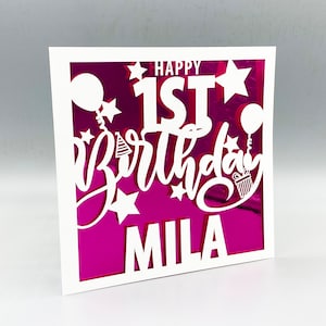 May include: White card with a pink background and a white cutout design. The design says "Happy 1st Birthday" with stars and balloons. The name "Mila" is at the bottom.