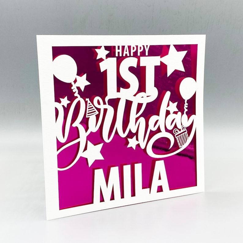May include: White card with a pink background and a white cutout design. The design says "Happy 1st Birthday" with stars and balloons. The name "Mila" is at the bottom.