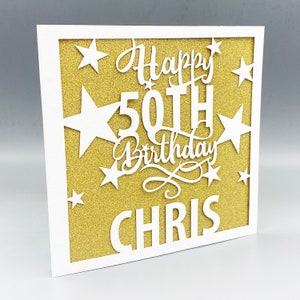 May include: A white card with a gold glitter background. The card has a white cut-out design that says "Happy 50th Birthday Chris" with stars around the text.
