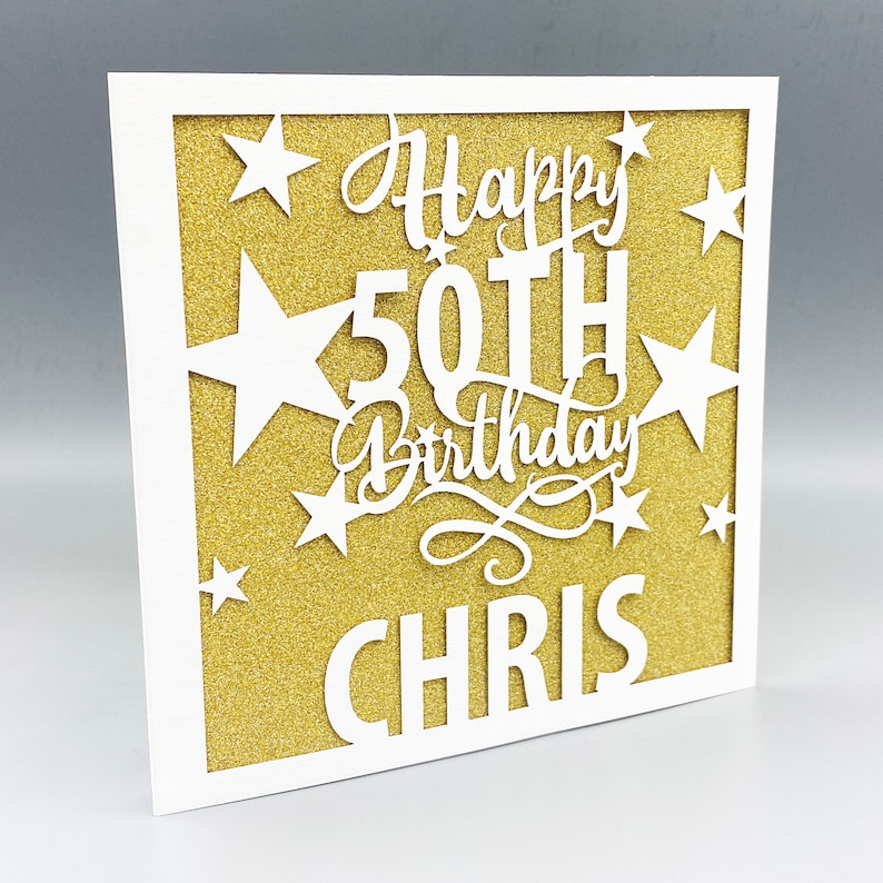 May include: A white card with a gold glitter background. The card has a white cut-out design that says "Happy 50th Birthday Chris" with stars around the text.