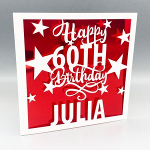 May include: A white card with a red background and white cut-out stars. The card says "Happy 60th Birthday Julia" in white lettering.
