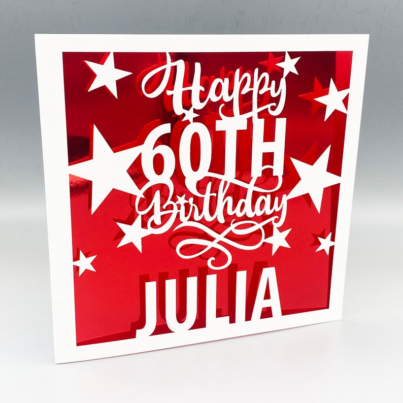 May include: A white card with a red background and white cut-out stars. The card says "Happy 60th Birthday Julia" in white lettering.