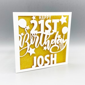 May include: White card with a gold glitter background. The card has a white cut-out design that says "Happy 21st Birthday Josh" with stars, balloons, and a party hat.