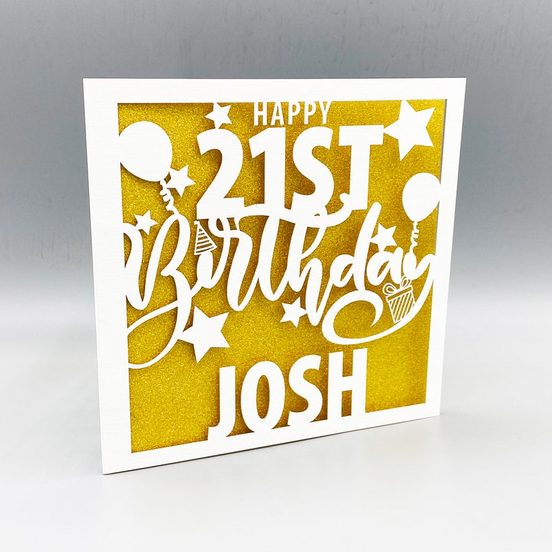 May include: White card with a gold glitter background. The card has a white cut-out design that says "Happy 21st Birthday Josh" with stars, balloons, and a party hat.
